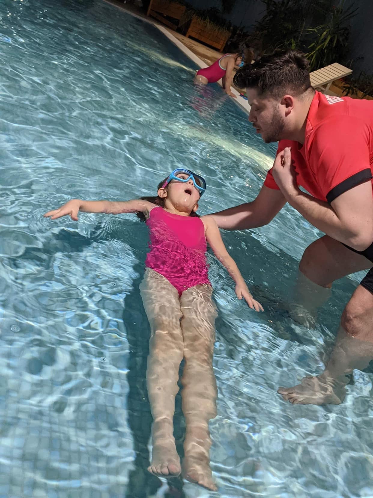 Instruktor w czerwonej koszulce asekuruje dziewczynkę w różowym stroju i niebieskich okularach, unoszącą się na wodzie w basenie z mozaiką, podczas lekcji.