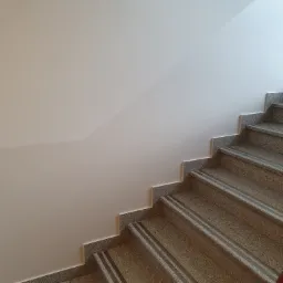 Klatka schodowa z granitowymi stopniami i białą ścianą, widok z góry na schody z antypoślizgowymi paskami.