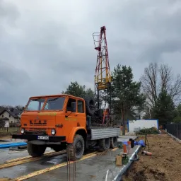 Pomarańczowy samochód ciężarowy STAR z masztem wiertniczym na placu budowy z widocznymi elementami fundamentów i rurami, pochmurne niebo w tle.