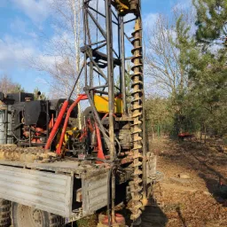 Wiertnica na podwoziu samochodowym z zamontowanym świdrem wiertniczym podczas pracy na zewnątrz, w tle drzewa i niebo.