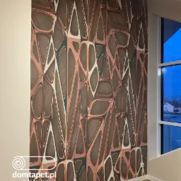 Ściana z nowoczesną tapetą o geometrycznym wzorze w odcieniach brązu, miedzi i bieli, z widocznym fragmentem okna i krajobrazem za nim.