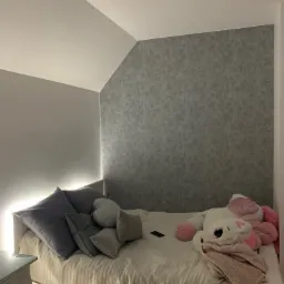 Pokój na poddaszu z szarą tapetą imitującą beton, oświetlenie LED za zagłówkiem łóżka, białe meble i pluszowy jednorożec na pościeli.