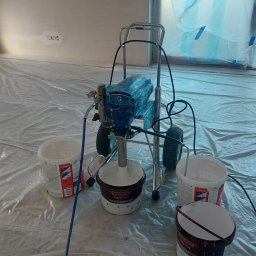 Firma Remontowo Wykończeniowa Michał Wójcik - Agregat malarski na wózku z pojemnikami farby gruntującej, zabezpieczona folią podłoga i okno, przygotowanie do malowania natryskowego.