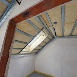 Firma Remontowo Wykończeniowa Michał Wójcik - Skośny sufit poddasza z oknem dachowym w trakcie prac: widoczne metalowe profile, izolacja z wełny mineralnej, fragment ściany z surowej cegły i częściowo otynkowane ściany.