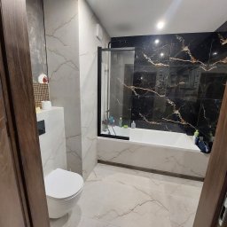 Firma Remontowo Wykończeniowa Michał Wójcik - Nowoczesna łazienka z czarno-złotą ścianą prysznicową, białą armaturą i płytkami imitującymi marmur, widziana zza częściowo otwartych, ciemnych drewnianych drzwi.