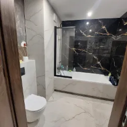 Nowoczesna łazienka z czarno-złotą ścianą prysznicową, białą armaturą i płytkami imitującymi marmur, widziana zza częściowo otwartych, ciemnych drewnianych drzwi.