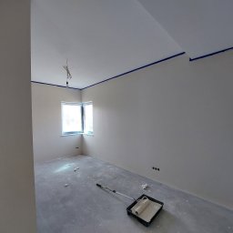 Firma Remontowo Wykończeniowa Michał Wójcik - Pokój po szpachlowaniu i gruntowaniu, przygotowany do malowania, z zabezpieczonymi taśmą narożnikami sufitu i oknem, widoczny wałek z farbą w kuwecie na podłodze.
