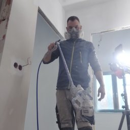 Firma Remontowo Wykończeniowa Michał Wójcik - Pracownik w masce ochronnej aplikuje gładź natryskową na ścianę w nowym mieszkaniu, widoczne gniazdka elektryczne, przewód i okno w tle.