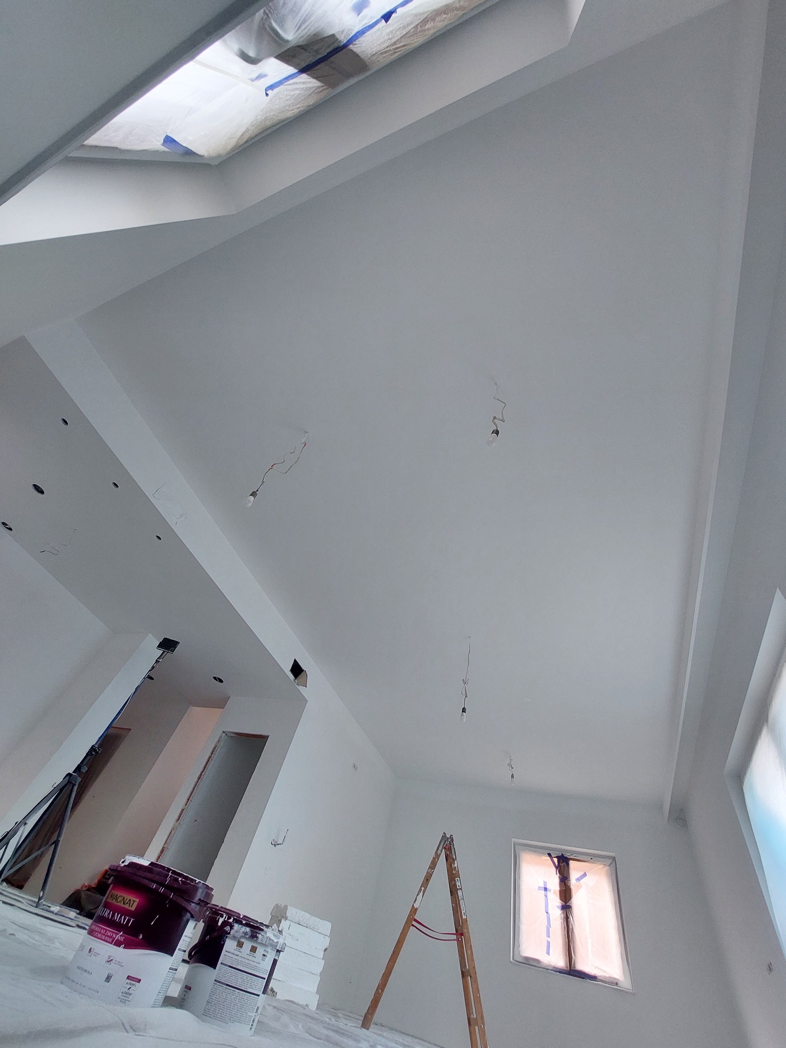Wnętrze w trakcie prac wykończeniowych: białe ściany i sufit, zabezpieczone okno, puszki z farbą oraz drabina