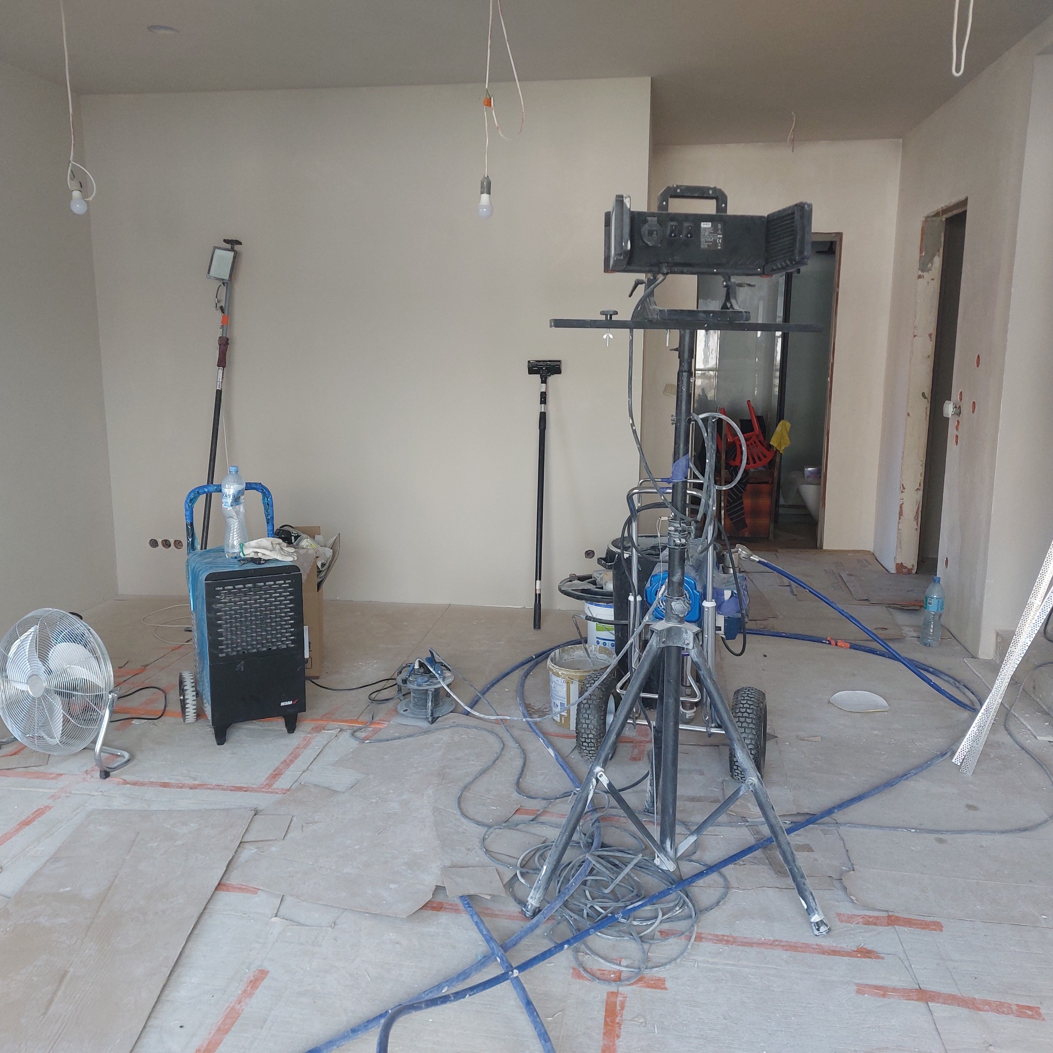 Wnętrze pokoju w trakcie malowania natryskowego, widoczny agregat malarski na statywie, wentylator, osuszacz powietrza, podłoga zabezpieczona kartonem, surowe żarówki zwisające z sufitu.