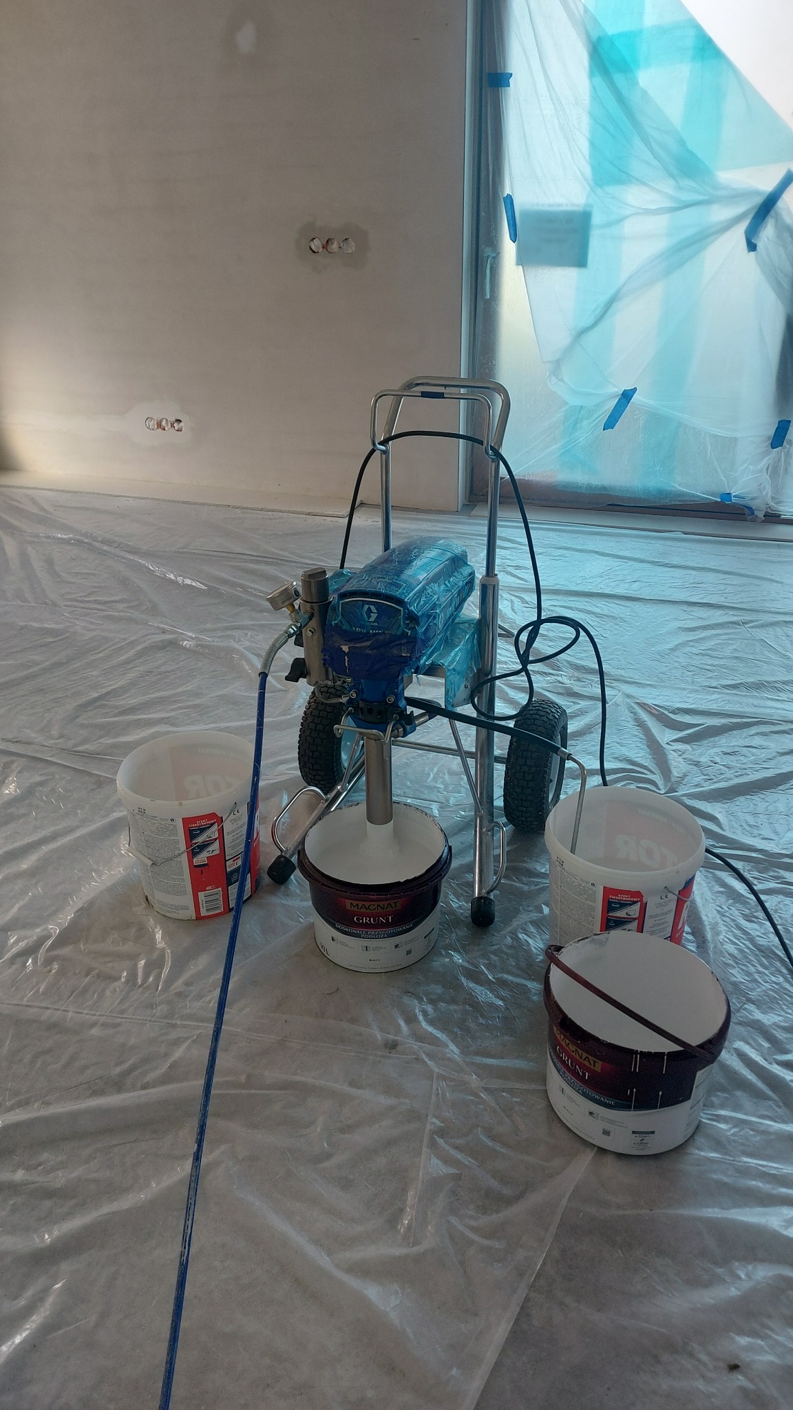 Agregat malarski na wózku z pojemnikami farby gruntującej, zabezpieczona folią podłoga i okno, przygotowanie do malowania natryskowego.