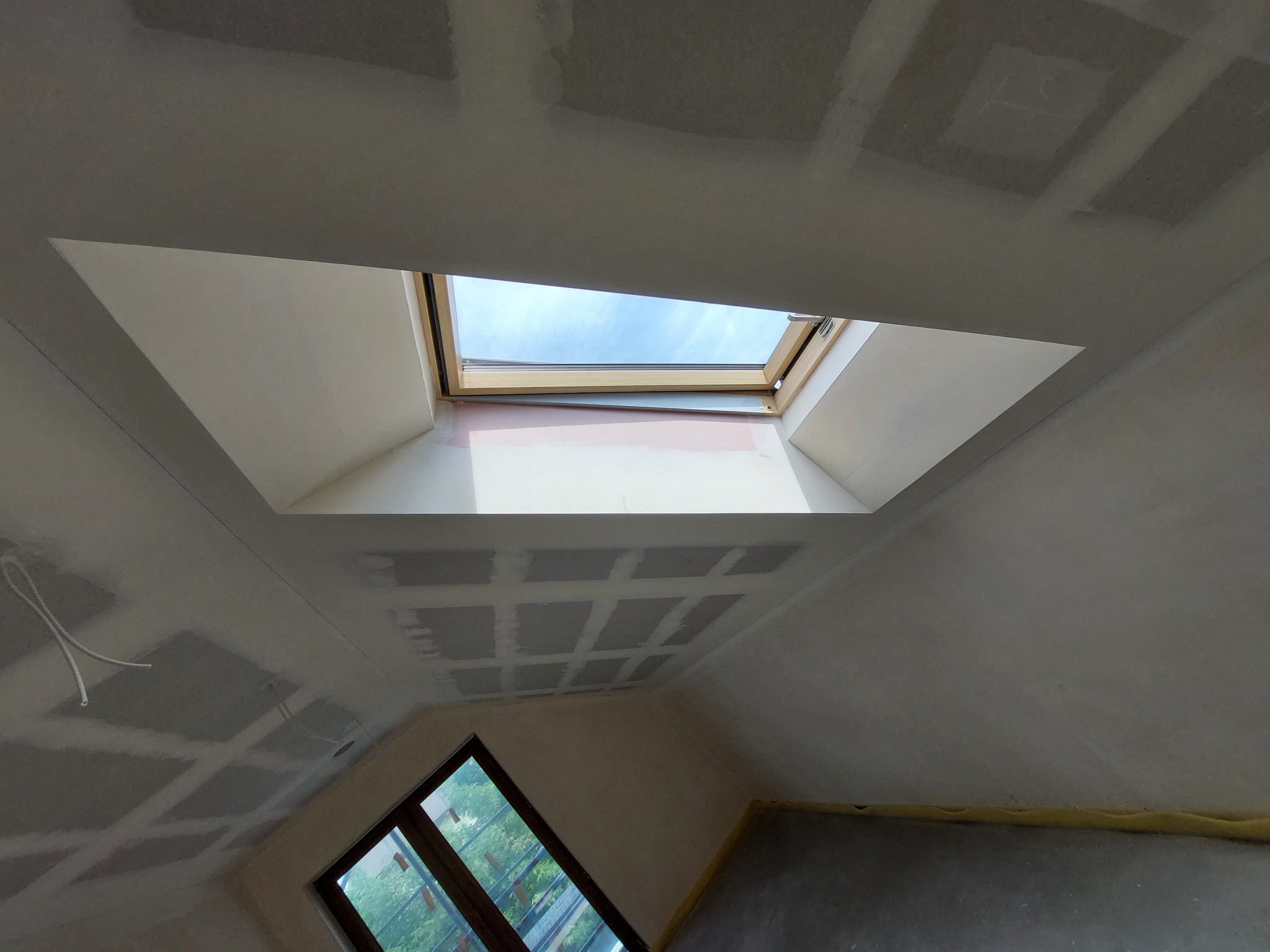 Sufit pod skosem w Katowicach z oknem dachowym i widocznymi łączeniami płyt karton-gips przed szpachlowaniem i malowaniem. Widoczne również okno fasadowe i fragment zabezpieczonej podłogi.