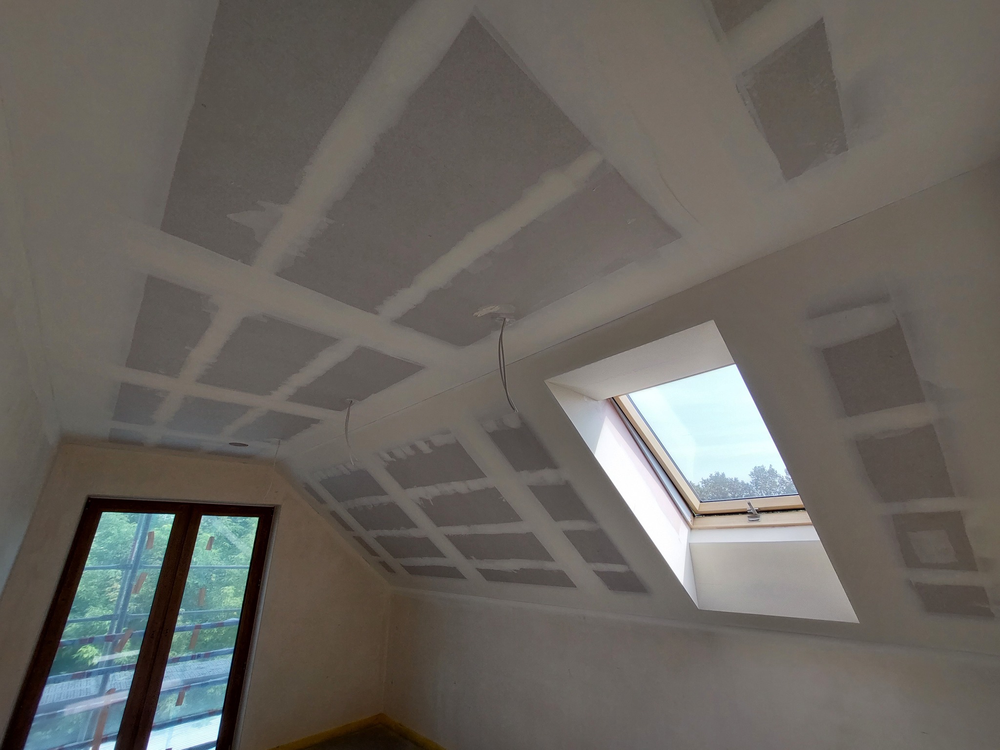 Poddasze w trakcie prac wykończeniowych: widoczne płyty kartonowo-gipsowe na skośnym suficie, spoiny zaszpachlowane, okno dachowe z widokiem na zieleń za oknem, oraz okno pionowe z widocznym...