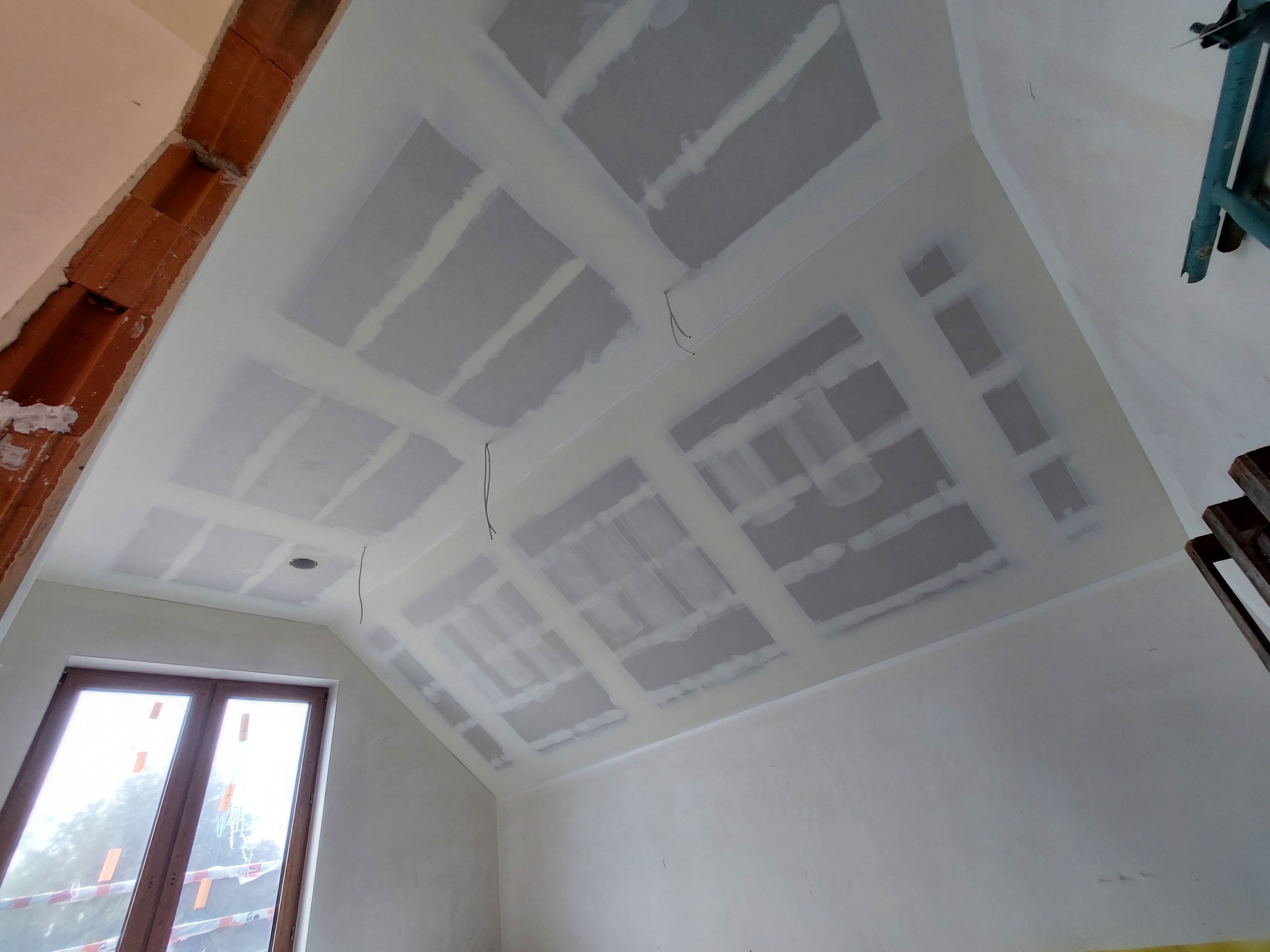 Skośny sufit poddasza z zamontowaną płytą karton-gips, widoczne łączenia płyt zabezpieczone taśmą i szpachlą, przygotowane do dalszych prac wykończeniowych, okno w tle.