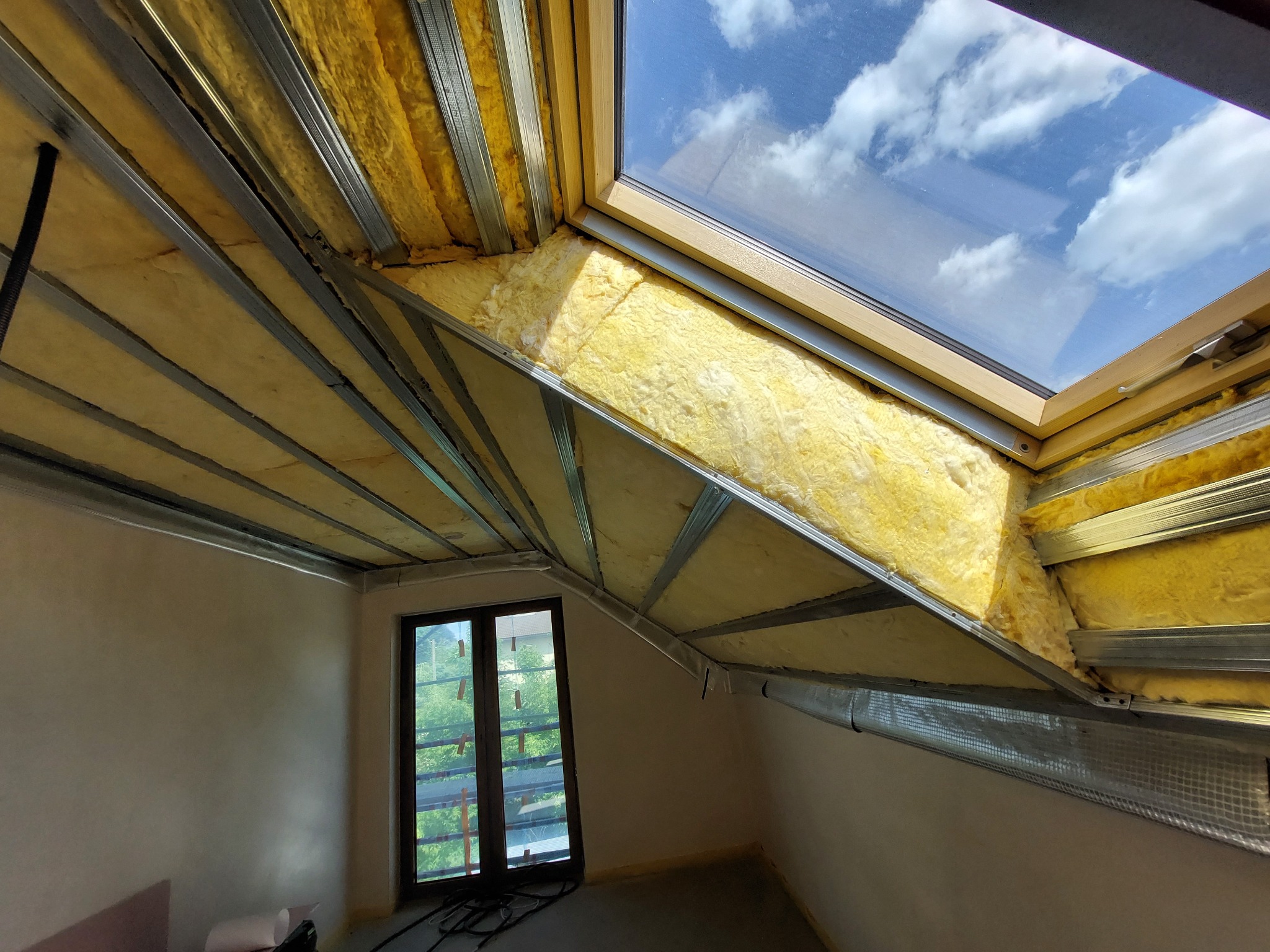Montaż wełny mineralnej między metalowymi profilami na poddaszu z oknem dachowym, widoczne fragmenty nieotynkowanej ściany i okno pionowe.