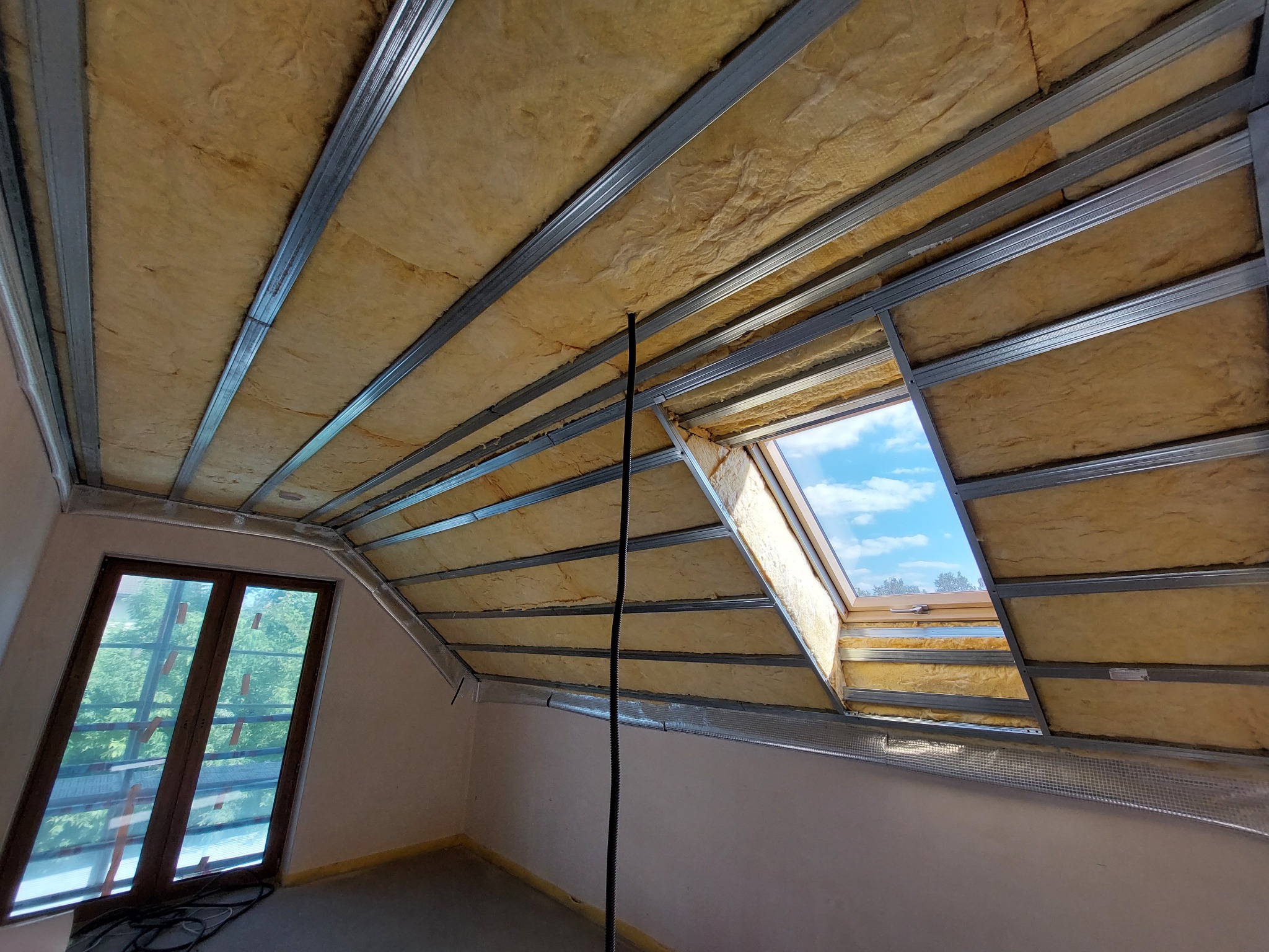 Poddasze w trakcie ocieplania wełną mineralną, widoczne metalowe profile konstrukcyjne i okno dachowe z widokiem na błękitne niebo.