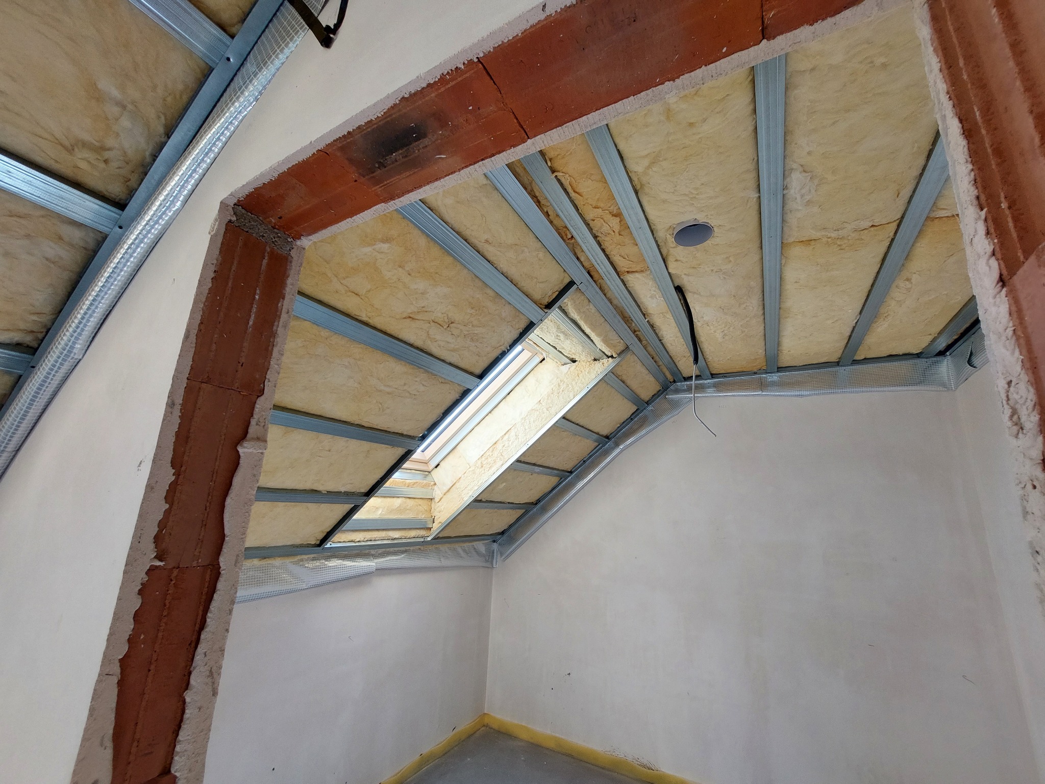 Skośny sufit poddasza z oknem dachowym w trakcie prac: widoczne metalowe profile, izolacja z wełny mineralnej, fragment ściany z surowej cegły i częściowo otynkowane ściany.