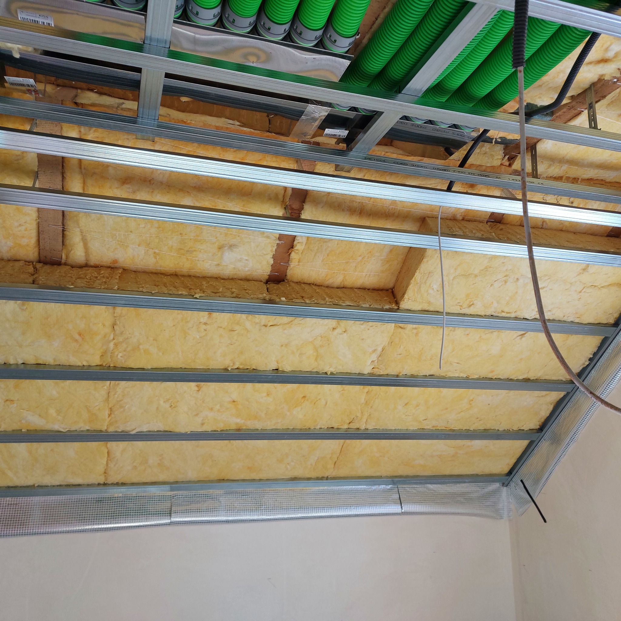 Montaż izolacji z wełny mineralnej w suficie podwieszanym z widoczną konstrukcją metalową i instalacją elektryczną w zielonych rurach karbowanych.