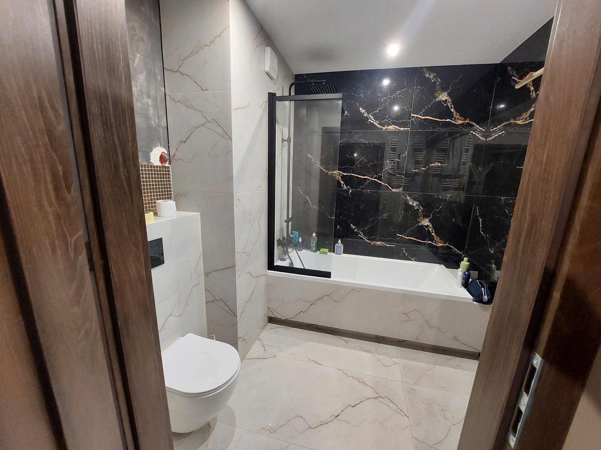 Nowoczesna łazienka z czarno-złotą ścianą prysznicową, białą armaturą i płytkami imitującymi marmur, widziana zza częściowo otwartych, ciemnych drewnianych drzwi.