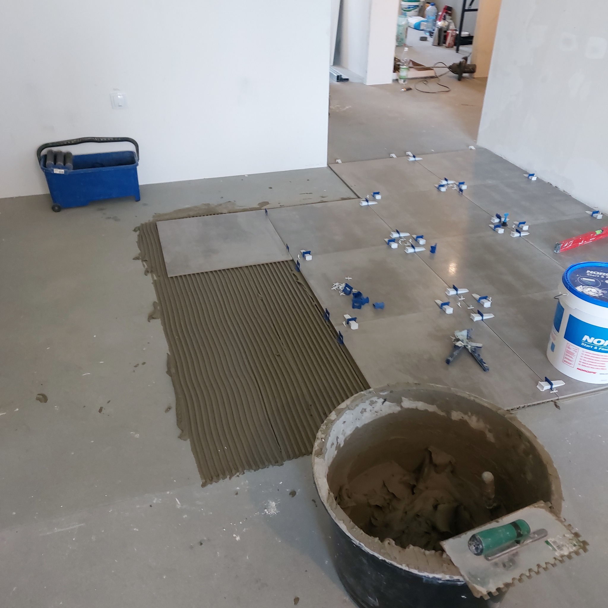 Układanie szarych płytek podłogowych z krzyżykami dystansowymi; widoczny klej na podłożu, wiadro z zaprawą i paca zębata.