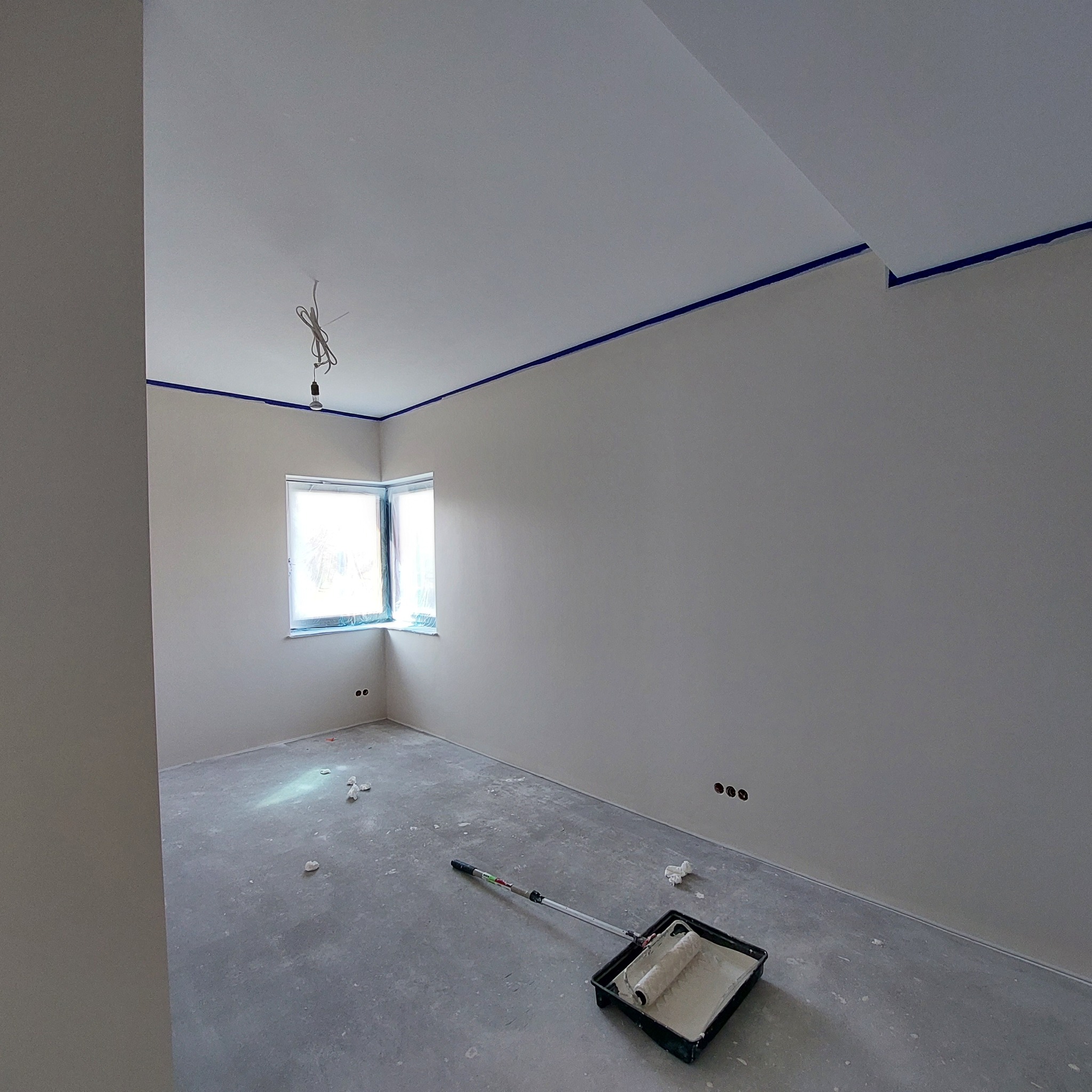 Pokój po szpachlowaniu i gruntowaniu, przygotowany do malowania, z zabezpieczonymi taśmą narożnikami sufitu i oknem, widoczny wałek z farbą w kuwecie na podłodze.
