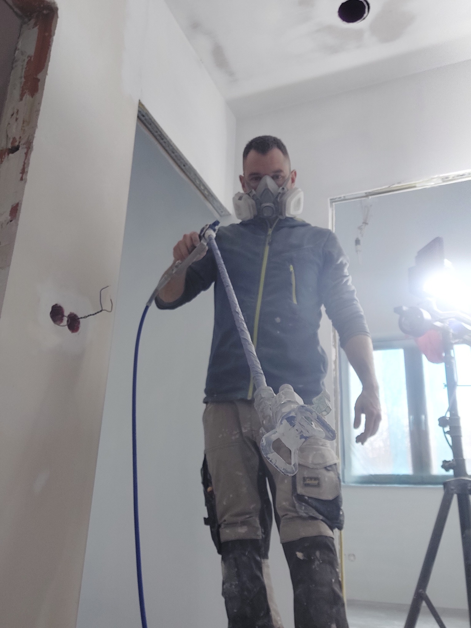 Pracownik w masce ochronnej aplikuje gładź natryskową na ścianę w nowym mieszkaniu, widoczne gniazdka elektryczne, przewód i okno w tle.