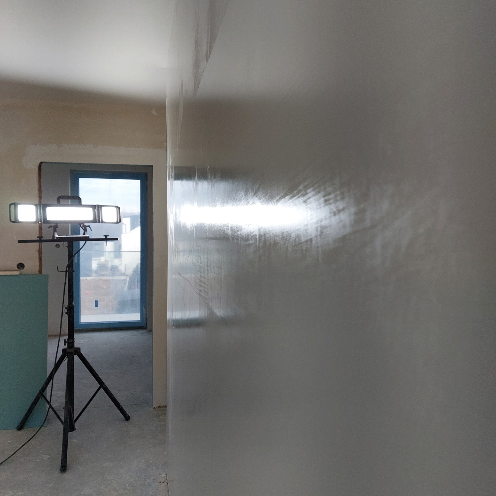 Świeżo nałożona, mokra gładź na ścianie w trakcie schnięcia, oświetlona lampą budowlaną na trójnogu, widoczne nierówności i ślady po szpachli.