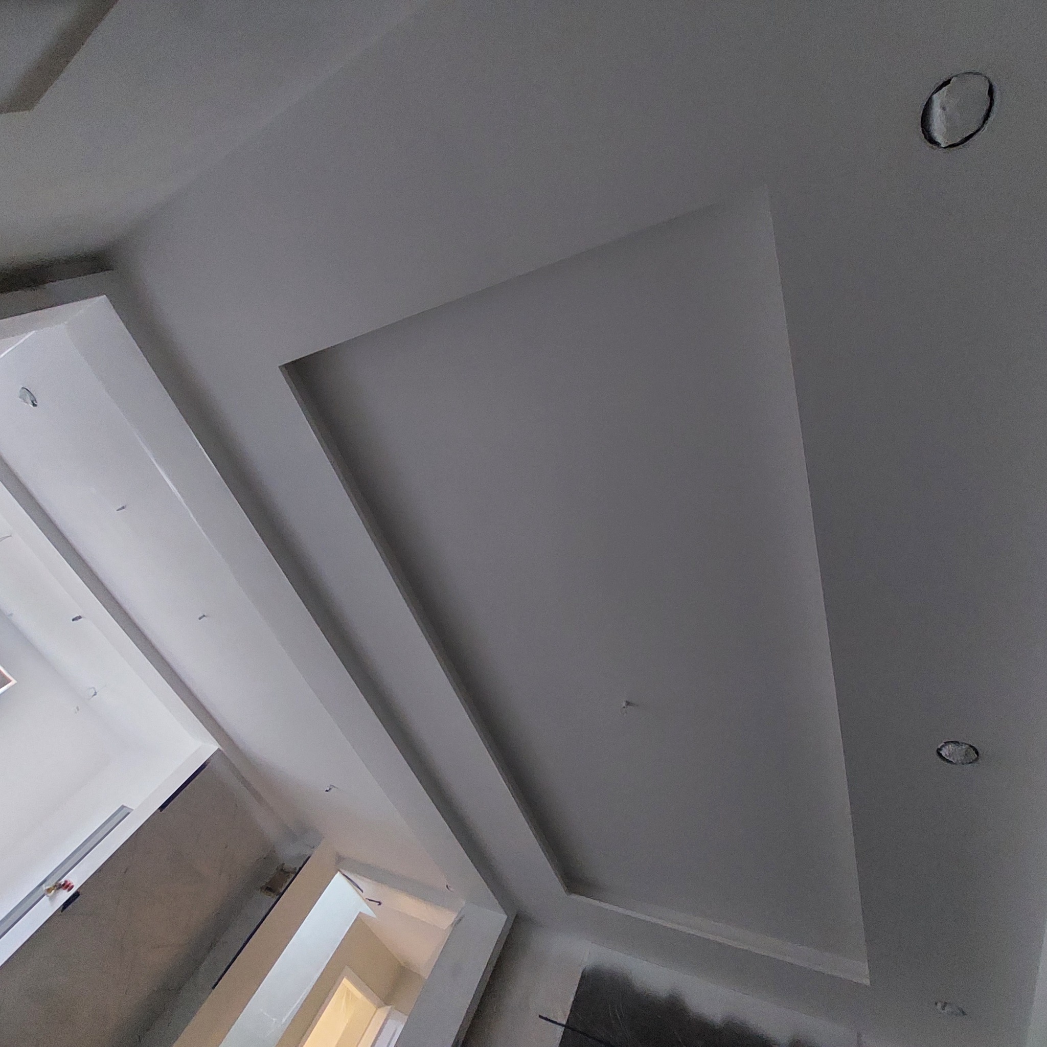 Biały sufit z wnęką i przygotowanymi otworami na oświetlenie, widok z dołu, fragment ściany w tle.