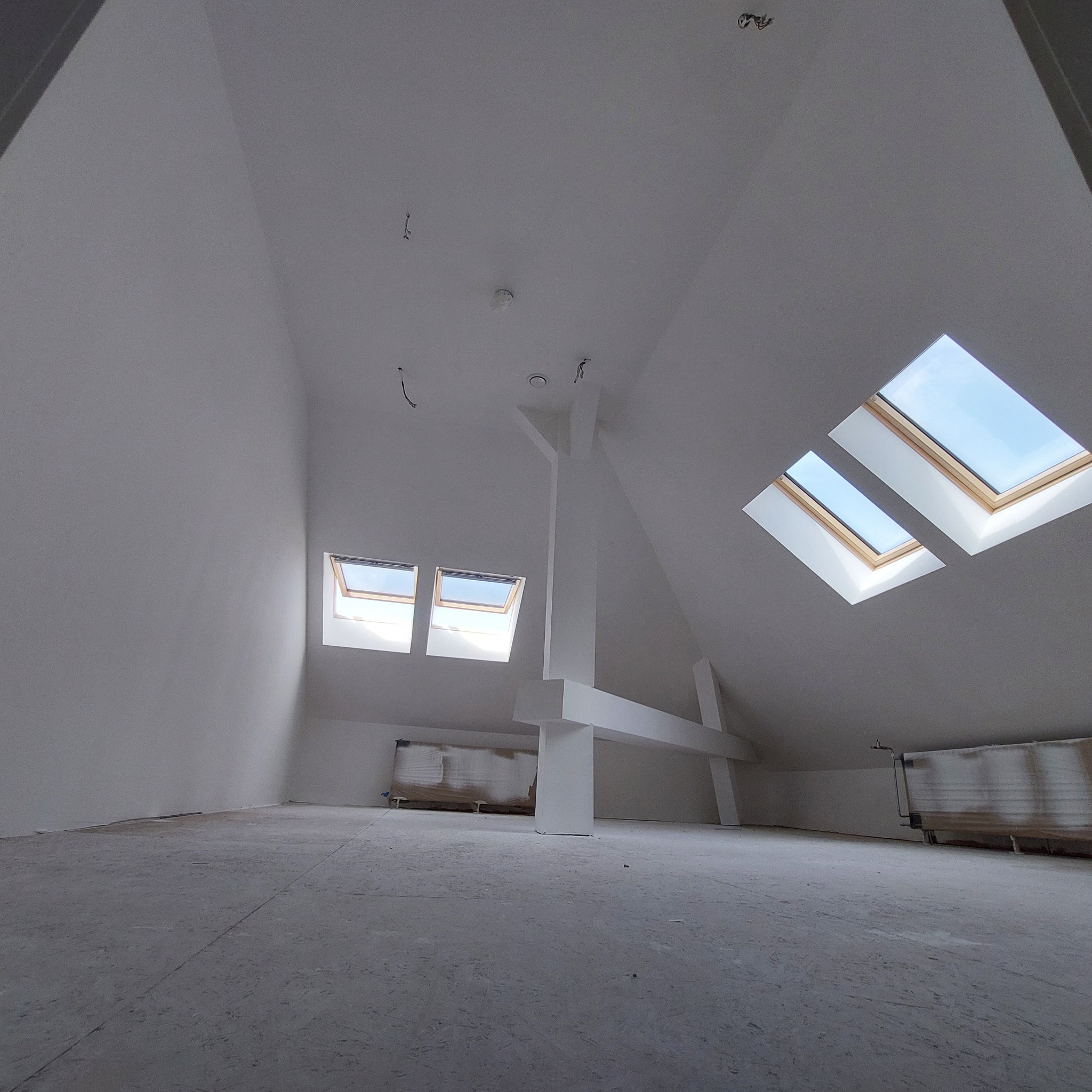 Poddasze po pracach wykończeniowych, świeżo pomalowane na biało, z widocznymi oknami dachowymi i odsłoniętymi grzejnikami, przygotowane do dalszego etapu aranżacji.