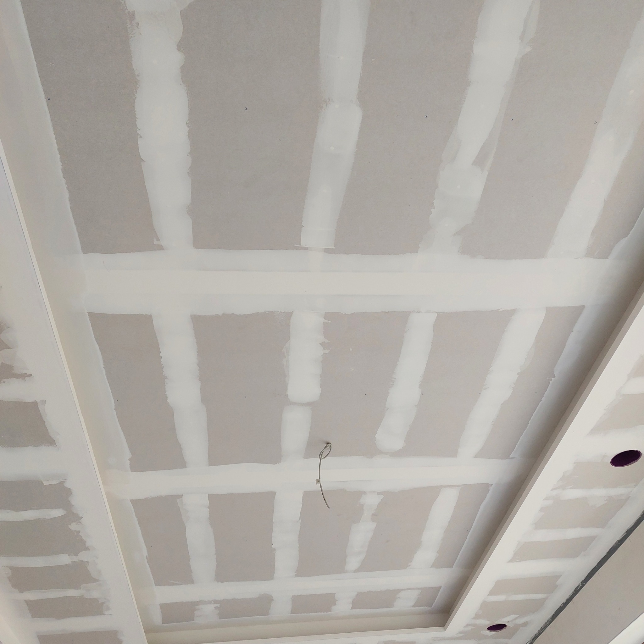 Sufit z płyt kartonowo-gipsowych z widocznymi łączeniami pokrytymi masą szpachlową, przygotowany do dalszych prac wykończeniowych, z wystającym przewodem elektrycznym i otworami na oświetlenie.