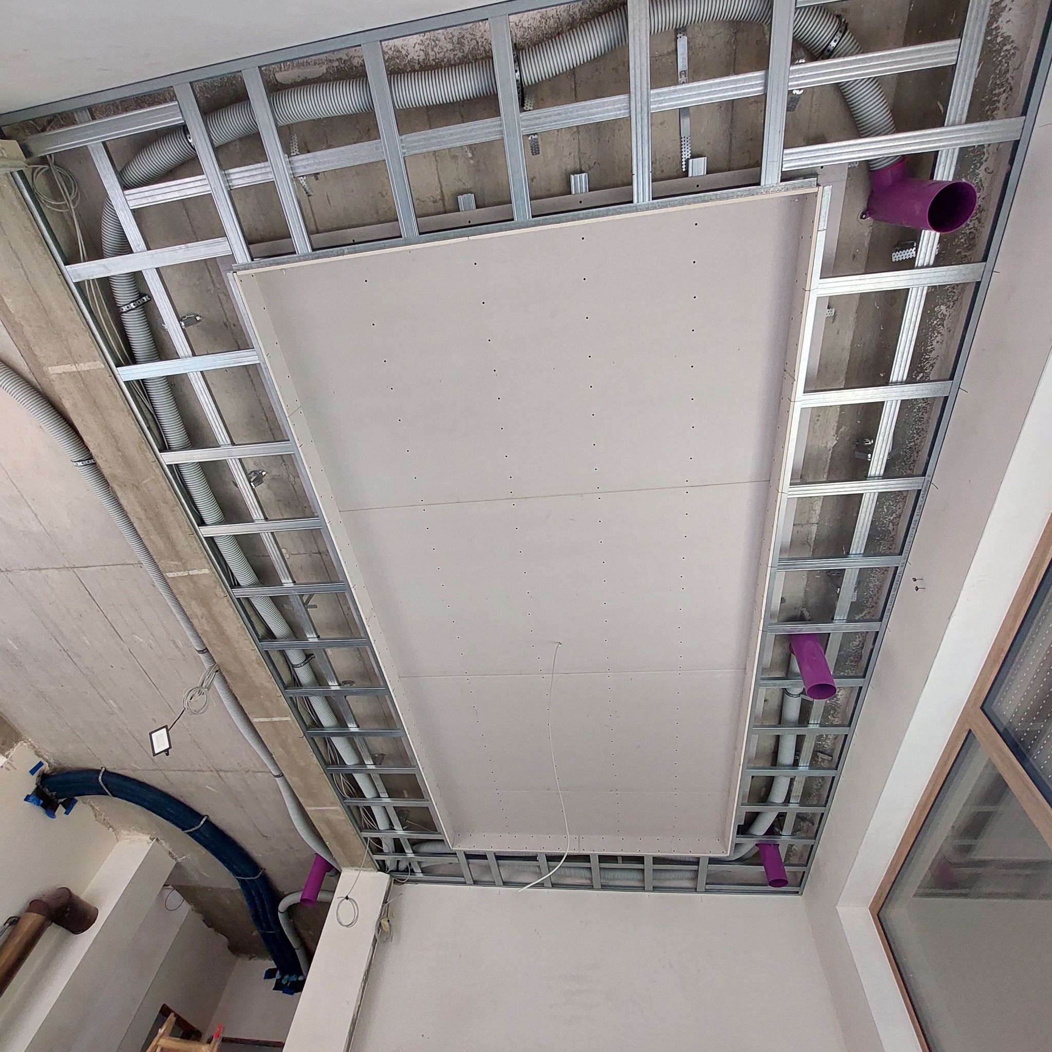 Metalowa konstrukcja sufitu podwieszanego z zamontowaną płytą kartonowo-gipsową, widoczne rury wentylacyjne i instalacje, fioletowe elementy kanalizacyjne.