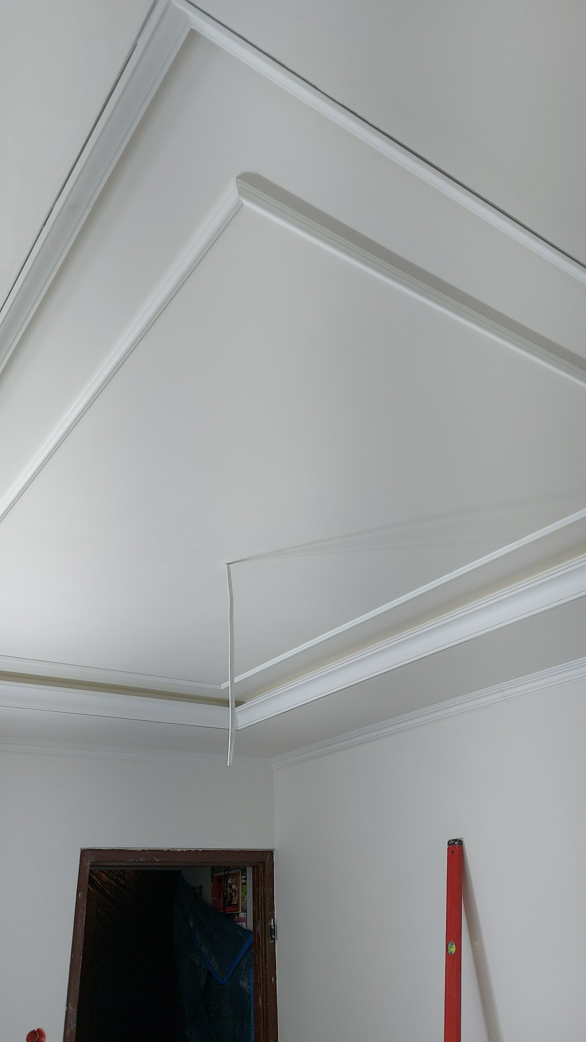 Biały sufit z dekoracyjnymi listwami przysufitowymi i kablem zwisającym z sufitu, widoczny fragment ściany z drzwiami i pionową poziomicą.