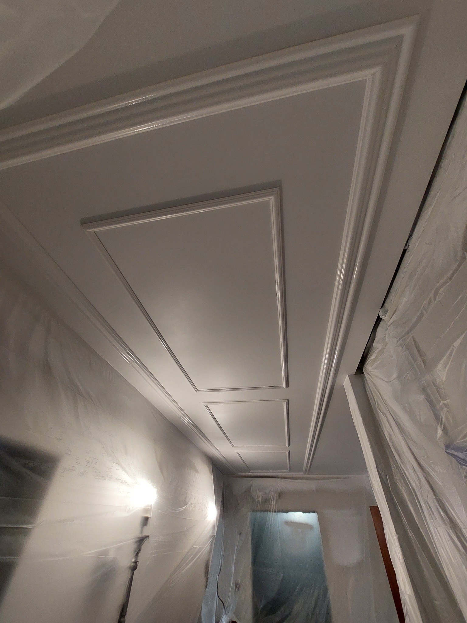 Biały sufit z dekoracyjnymi listwami w kształcie prostokątów, zabezpieczony folią malarską, widok z dołu.