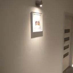 Ściana pomalowana na jasny kolor z zawieszonym obrazem w białej ramie, oświetlonym przez kinkiet, widoczny fragment drewnianych drzwi z przeszkleniami.