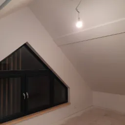 Poddasze ze skośnym sufitem i trójkątnym oknem z czarną ramą oraz drewnianym parapetem, surowy stan po malowaniu, widoczna żarówka na kablu.