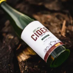 Butelka zielonego jabłkowego cydru leży ukośnie na ciemnym, teksturowanym tle przypominającym korę drzewa, z widoczną etykietą w odcieniach bieli i czerwieni.