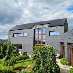 Dom jednorodzinny z szarej dachówki z panelami słonecznymi na dachu, widok z ogrodu w słoneczny dzień.