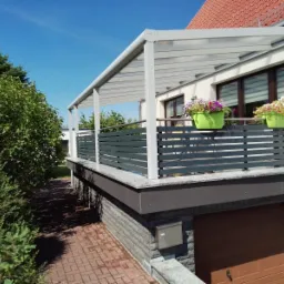 Zadaszony balkon z szarą, poziomą balustradą i dwoma zielonymi donicami z kwiatami, widok z zewnątrz na tle błękitnego nieba i zieleni.