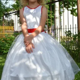 Dziewczynka w białej sukience z czerwonymi akcentami, stojąca na trawie przed ogrodzeniem.