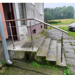 Piotr Sroka uslugi spawalnicze - Zewnętrzne schody z metalową, minimalistyczną balustradą ze stali nierdzewnej przy budynku z szarą elewacją. Widoczne otoczenie budynku i zaparkowany samochód.