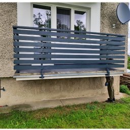 Piotr Sroka uslugi spawalnicze - Szara, pozioma balustrada balkonowa z metalu, zamontowana na elewacji domu. Na balkonie płytki, w tle okno i antena satelitarna. Widoczny fragment osoby w rękawicach.