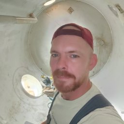 Piotr Sroka uslugi spawalnicze - Spawacz wewnątrz metalowej rury, widoczne narzędzia i otwór oświetleniowy,  w burgundowej czapce z daszkiem,  spogląda w obiektyw.