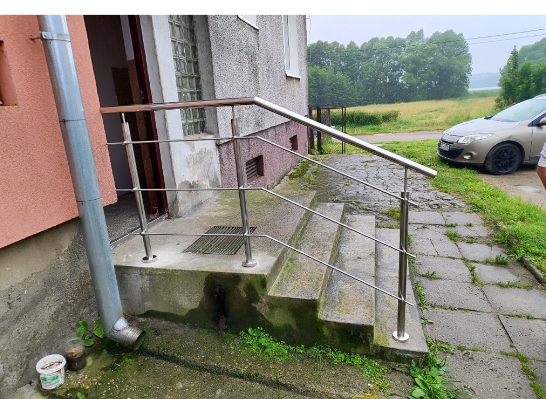 Zewnętrzne schody z metalową, minimalistyczną balustradą ze stali nierdzewnej przy budynku z szarą elewacją. Widoczne otoczenie budynku i zaparkowany samochód.
