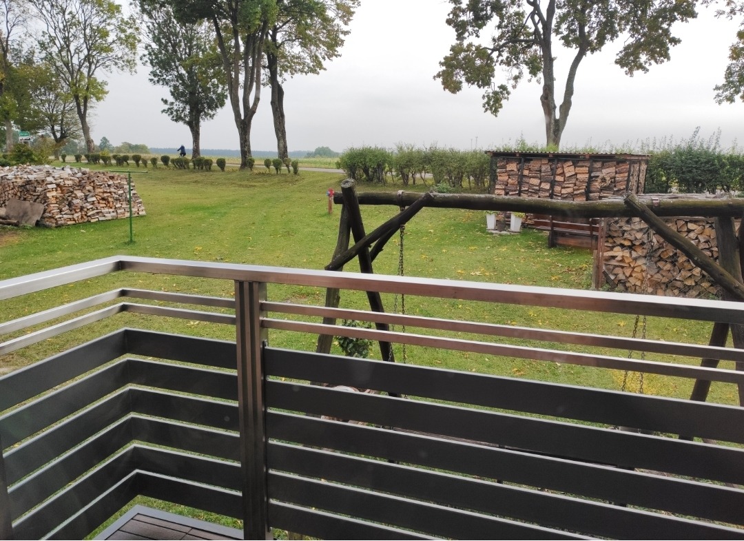 Nowoczesna balustrada ze stali nierdzewnej i ciemnego drewna na tle zielonego ogrodu z huśtawką i stertą drewna. Ujęcie z niskiego kąta.