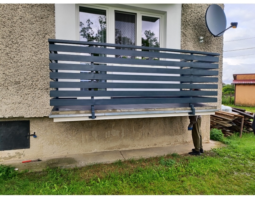 Szara, pozioma balustrada balkonowa z metalu, zamontowana na elewacji domu. Na balkonie płytki, w tle okno i antena satelitarna. Widoczny fragment osoby w rękawicach.