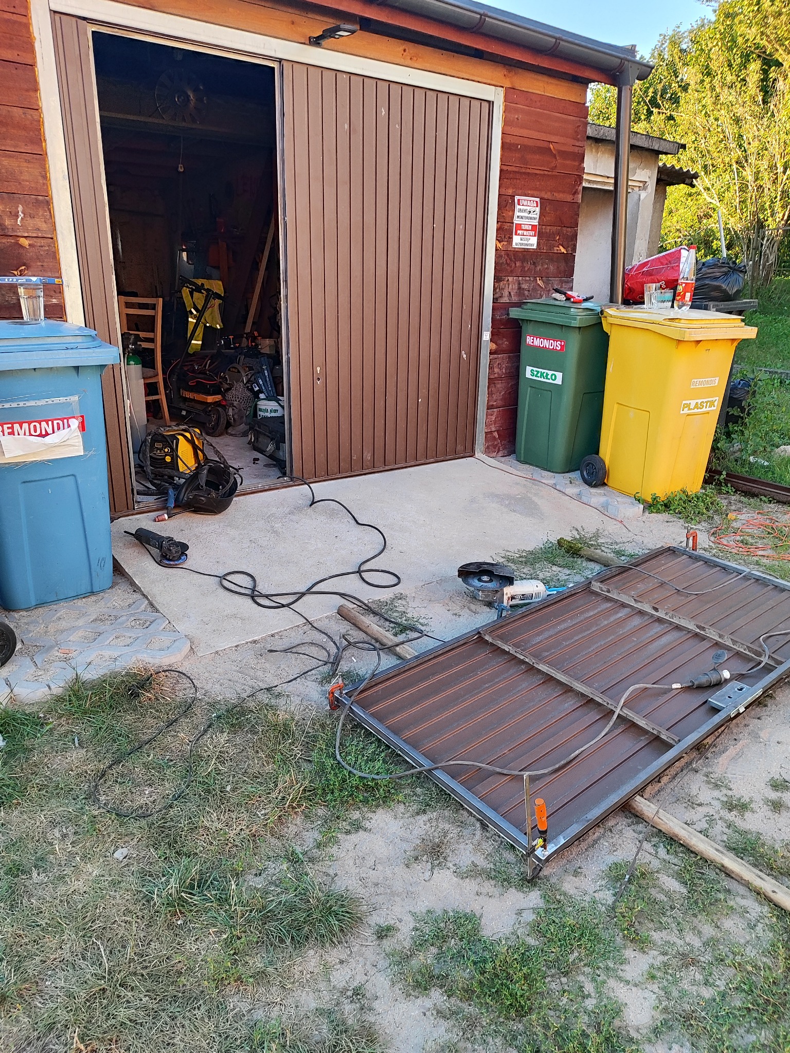 Brązowa brama garażowa z otwartymi drzwiami, obok narzędzia ślusarskie i spawarka. Na pierwszym planie rama bramy w trakcie obróbki, na zewnątrz budynku.