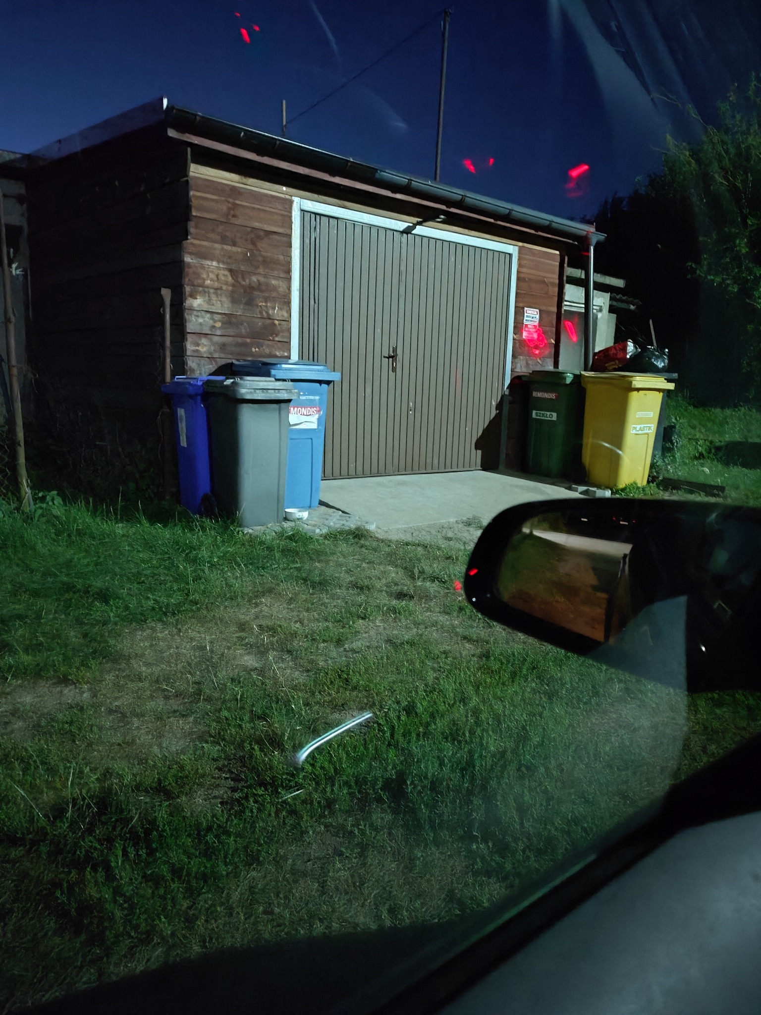 Drewniany garaż z metalowymi drzwiami na posesji, obok stoją pojemniki na odpady w różnych kolorach, widok z wnętrza pojazdu.