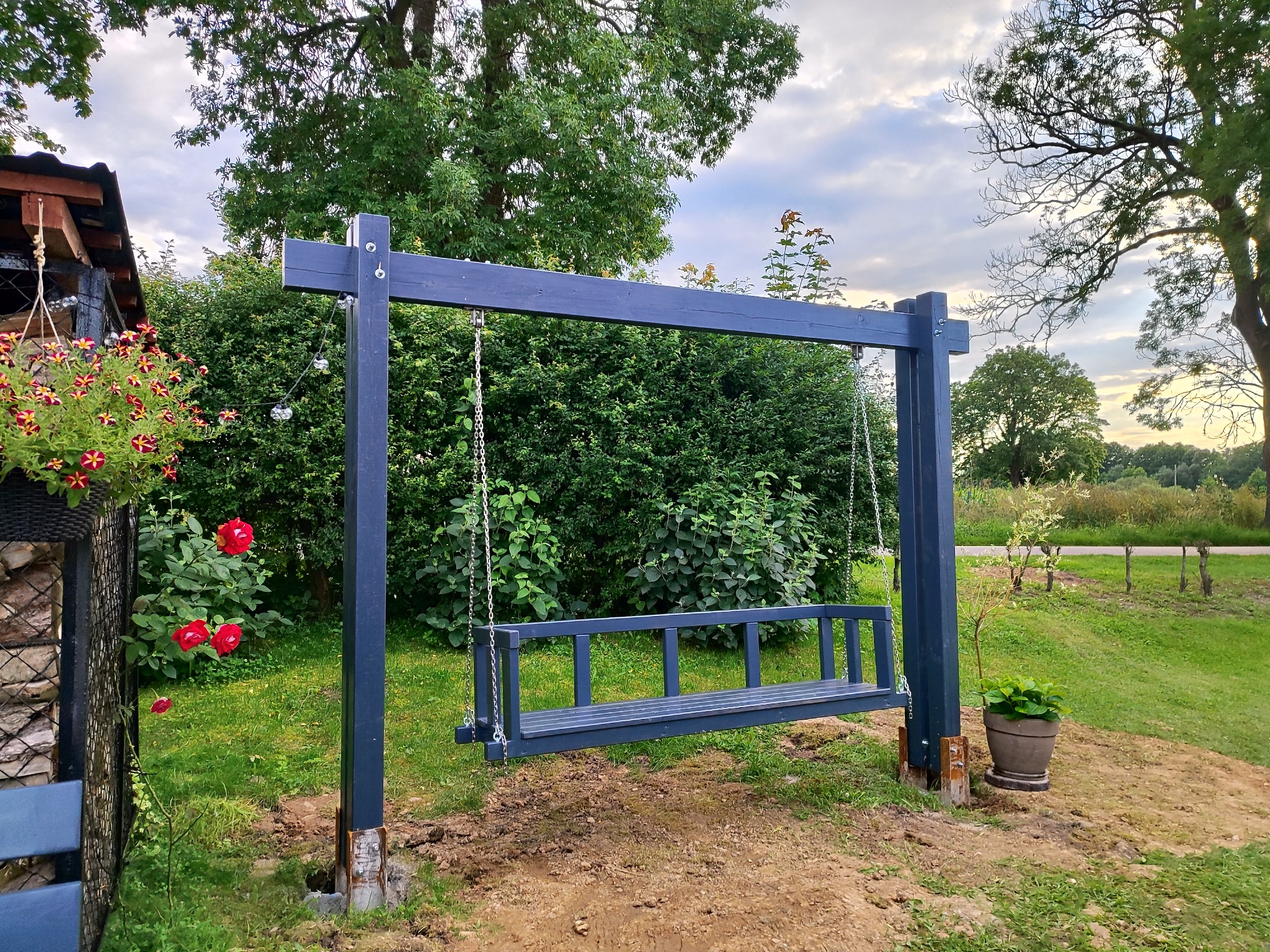Nowoczesna, ciemnoniebieska huśtawka ogrodowa na łańcuchach, z drewnianą konstrukcją, na tle zieleni i kwiatów. Elementy dekoracyjne: donica i wiszące kwiaty.