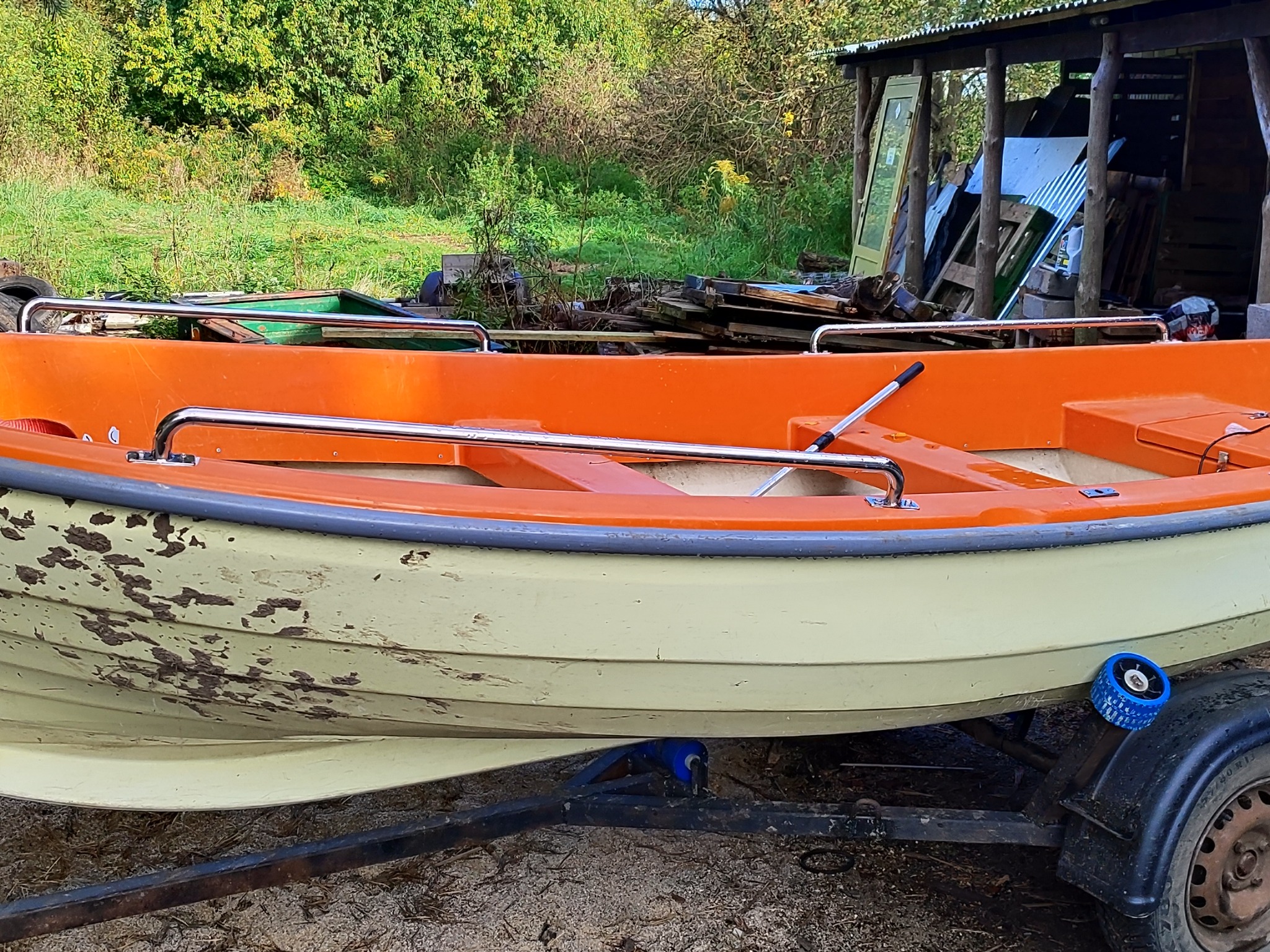 Pomarańczowo-żółta łódź z chromowanymi poręczami na przyczepie, na tle zieleni i drewnianej konstrukcji. Widoczne ślady użytkowania na kadłubie łodzi.