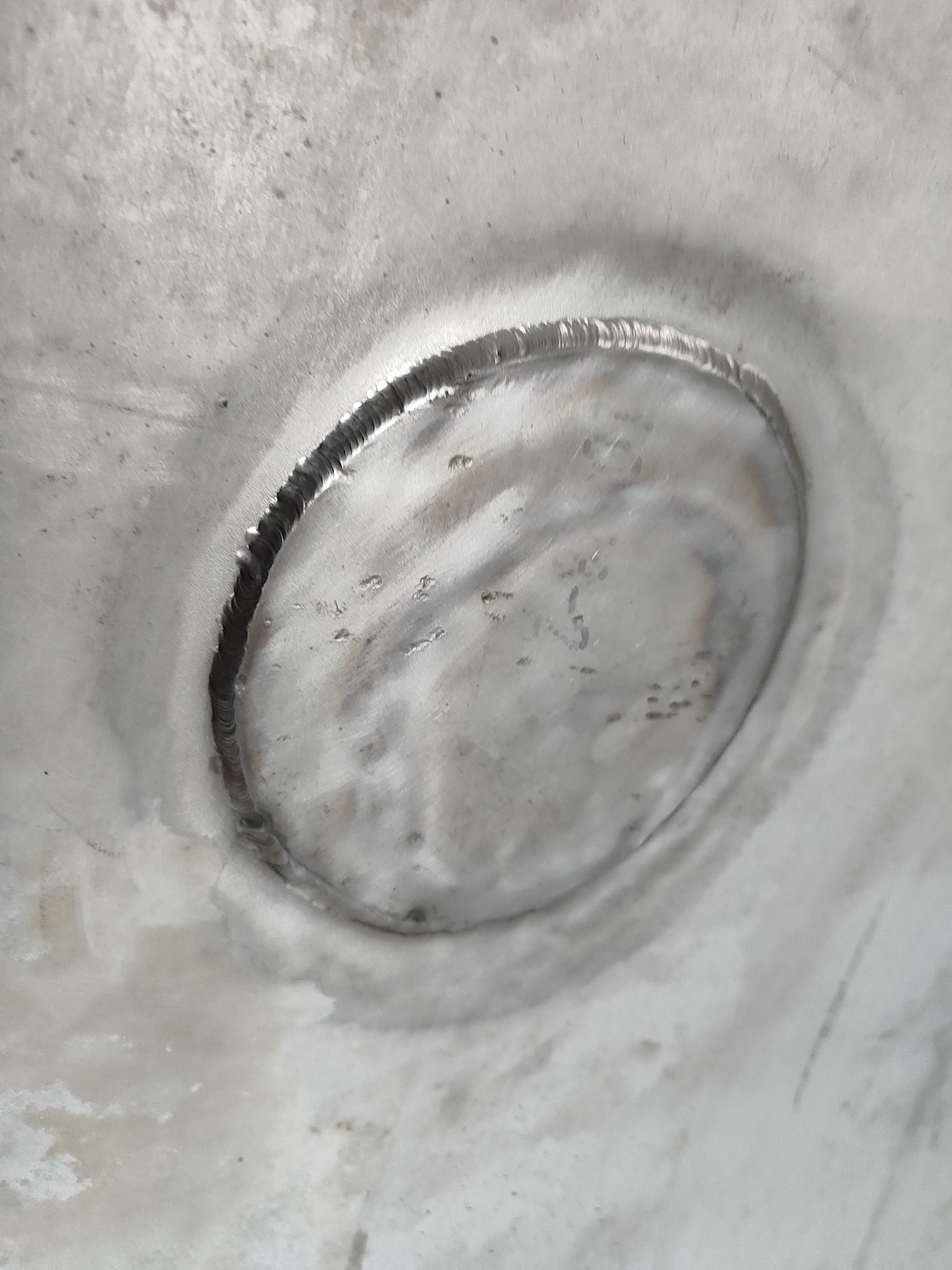 Zbliżenie na spoinę okrągłą na metalowej powierzchni, widoczne ślady po spawaniu, szary odcień metalu, delikatne nierówności.